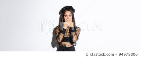 Shocked woman in witch outfit gasping and cover mouth speechless, standing over white background, looking at something surprising, wearing black lace dress and wreath, white background 94978800