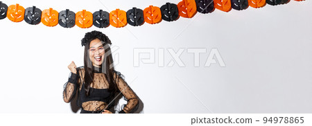Image of happy asian woman in witch costume having fun on halloween party, saying yes cheerful, standing against pumpkin banners Image of happy asian woman in witch costume having fun on halloween party, saying yes cheerful, standing against pumpkin banners 94978865