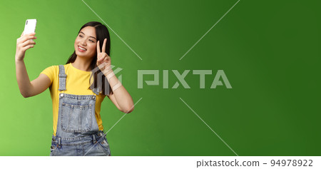 Tender cute asian brunette female, hold smartphone, posing selfie, look telephone screen, photographing, show victory peace sign, joyfully chatting friend abroad, stand green background Tender cute asian brunette female, hold smartphone, posing selfie, look telephone screen, photographing, show victory peace sign, joyfully chatting friend abroad, stand green background 94978922