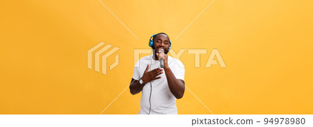 Portrait of cheerful positive chic handsome african man holding microphone and having headphones on head listening music singing song enjoying weekend vacation isolated on yellow background 94978980