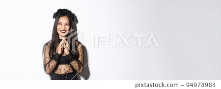 Image of beautiful asian woman dressed-up as a witch or widow in black lace dress, gothic wreath, smiling pleased upper left corner and steeple fingers while prepare plan Image of beautiful asian woman dressed-up as a witch or widow in black lace dress, gothic wreath, smiling pleased upper left corner and steeple fingers while prepare plan 94978983