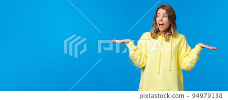 Girl weighing two choices as making decision, look away picking between products she holding in arms on left and right side, standing excited over blue background Girl weighing two choices as making decision, look away picking between products she holding in arms on left and right side, standing excited over blue background 94979138