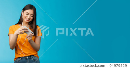 Cute entertained good-looking urban female student spend break playing racing game, hold smartphone horizontal tilt phone move car, smiling broadly standing enthusiastic blue background 94979329