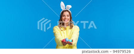 Holidays, people and fun concept. Charismatic cute blond girl holding coloured Easter eggs, wearing bunny ears and smiling joyfully, congratulate family with holy day, blue background Holidays, people and fun concept. Charismatic cute blond girl holding coloured Easter eggs, wearing bunny ears and smiling joyfully, congratulate family with holy day, blue background 94979526