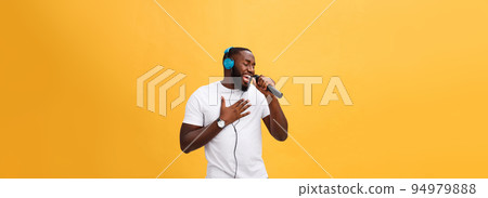 Portrait of cheerful positive chic handsome african man holding microphone and having headphones on head listening music singing song enjoying weekend vacation isolated on yellow background Portrait of cheerful positive chic handsome african man holding microphone and having headphones on head listening music singing song enjoying weekend vacation isolated on yellow background 94979888