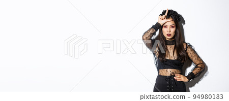Image of arrogant sexy asian woman in witch costume showing loser gesture on forehead, mocking someone at halloween party, standing over white background Image of arrogant sexy asian woman in witch costume showing loser gesture on forehead, mocking someone at halloween party, standing over white background 94980183
