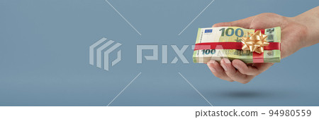 Gift of money. A man holds a stack of banknotes in his hands as a symbol or concept of a gift. 100 euro banknotes tied with a red ribbon with a bow on a blue background. Gift of money. A man holds a stack of banknotes in his hands as a symbol or concept of a gift. 100 euro banknotes tied with a red ribbon with a bow on a blue background. 94980559