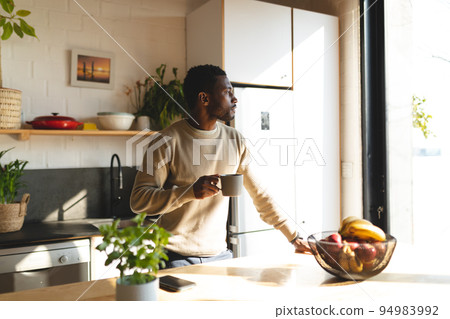 Happy african american man leaning on countertop and drinking coffee in kitchen Happy african american man leaning on countertop and drinking coffee in kitchen 94983992