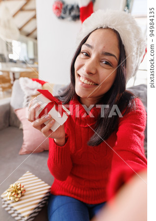Happy biracial woman wearing santa hat, sitting on sofa in living room, having video call 94984135