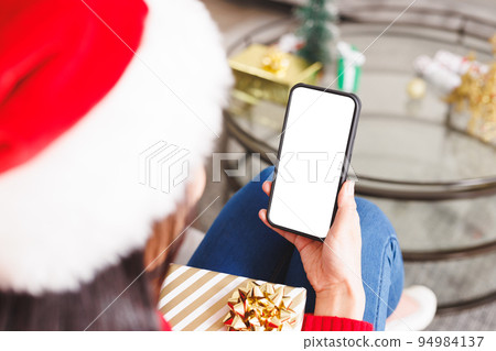 Biracial woman wearing santa hat, sitting on sofa in living room, using smartphone with copyspace Biracial woman wearing santa hat, sitting on sofa in living room, using smartphone with copyspace 94984137