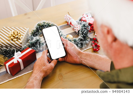 Caucasian man wearing santa hat, sitting at table in kitchen, using smartphone with copyspace 94984143