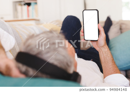 Caucasian man laying on sofa in living room and using smartphone with copyspace Caucasian man laying on sofa in living room and using smartphone with copyspace 94984148