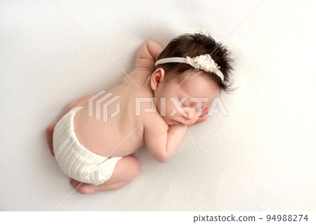 Top view of a newborn baby girl sleeping in white panties, with a white bandage her head on a white bed. Beautiful portrait of a little girl 7 days, one week. Professional photography in macro studio. Top view of a newborn baby girl sleeping in white panties, with a white bandage her head on a white bed. Beautiful portrait of a little girl 7 days, one week. Professional photography in macro studio. 94988274