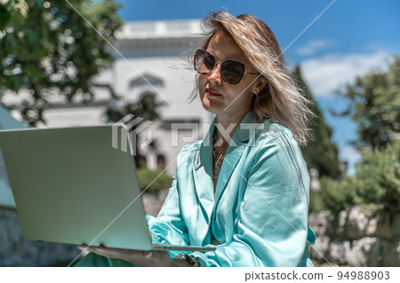 a young beautiful woman with blond curly hair in glasses and a blue dress sits on the grass in nature and uses a laptop, 94988903