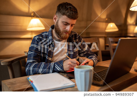 Casual business man or freelancer planning his work on notebook, working on laptop computer with smart phone, cup of coffee on table at coffee shop or home office. 94989307