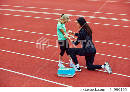 First aid for a young athlete at stadium. Sport teen girl suffering with pain of injury after running. Injury from workout concept. Concept of sport, fitness, achievements, studying First aid for a young athlete at stadium. Sport teen girl suffering with pain of injury after running. Injury from workout concept. Concept of sport, fitness, achievements, studying 94990163