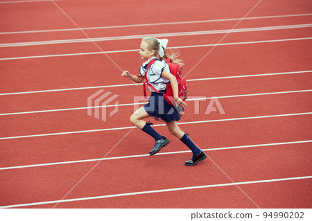 Start. Back to school, kids and education concept. Girl dressed in school uniform as elementary student carrying big backpack running on treadmill at the stadium or arena. 94990202