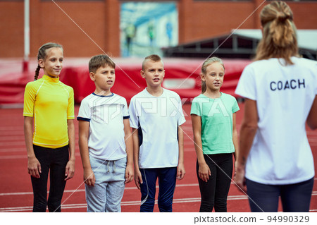 Female coach training athletes. Group of children before running on treadmill at the stadium. Concept of sport, achievements, studying, goals, skills. Little boys and girls training outdoor. 94990329