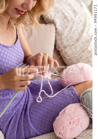Close-up of woman in a lilac home dress knits a light pink scarf or plaid from natural threads on the bed. 94991171
