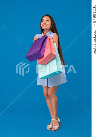 Beautiful young woman holding shopping bags and looking away while standing against blue background. 94991236
