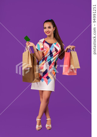 Attractive woman 20s in dress holding shopping packages and showing green credit card isolated over purple background 94991251