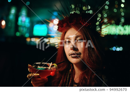 Female with skeleton makeup at a Halloween party holds cocktail and looks at camera. All Saints' Night. Celebration in a nightclub. Woman in creepy costume of Mexican goddess of death. Female with skeleton makeup at a Halloween party holds cocktail and looks at camera. All Saints' Night. Celebration in a nightclub. Woman in creepy costume of Mexican goddess of death. 94992670