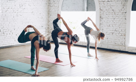 Three pretty young ladies are practicing Lord of the dance pose Natarajasana while doing yoga in modern wellness center with hign ceiling. Concentration flexibility energy concept. Three pretty young ladies are practicing Lord of the dance pose Natarajasana while doing yoga in modern wellness center with hign ceiling. Concentration flexibility energy concept. 94992786
