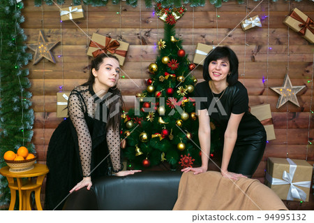 Mom with an adult daughter near a wooden wall with a decorated Christmas tree in anticipation of the new year Mom with an adult daughter near a wooden wall with a decorated Christmas tree in anticipation of the new year 94995132