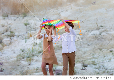 A girl and boy are carrying a bright kite with their hands raised high above their heads A girl and boy are carrying a bright kite with their hands raised high above their heads 94995137