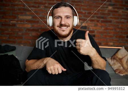 Handsome bearded man in headphones listening to music at home. Music. Half-length portrait of young guy in casual youth style clothes with sign ok sitting on sofa. Concept of emotions. Handsome bearded man in headphones listening to music at home. Music. Half-length portrait of young guy in casual youth style clothes with sign ok sitting on sofa. Concept of emotions. 94995812
