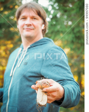 Satisfied young man holding a found porcini mushroom in his hands. The mushroom picker holds a cep mushroom. Man collect mushrooms in forest.  94997080