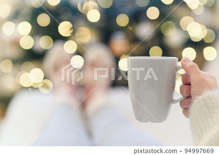 A woman with cup of coffee in woolen socks lying on the sofa near Christmas tree with garland 94997669