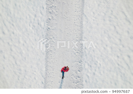 Woman in a red christmas jacket with backpack walking on the snow and doing footprints in a park. Winter. Drone, top, aerial view. 94997687
