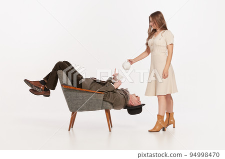 Creative portrait of stylish man in suit and cap sitting upside down on chair. Woman pouring tea into his cup 94998470