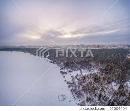 Snow-covered forest on lake shore with ice at sunset and the city on horizon, auerial view Snow-covered forest on lake shore with ice at sunset and the city on horizon, auerial view 95004404