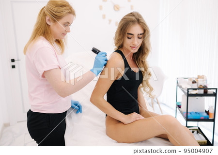 Female doctor dermatologist examines birthmarks and skin moles of the patient with a dermatoscope. 95004907