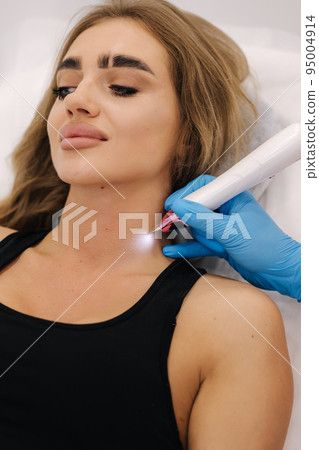 Plasma pen reducing neoplasm around neck on young woman. Woman at cosmetology clinic taking a beauty procedure for skin. Noninvasive cosmetic treatment. Plasma pen reducing neoplasm around neck on young woman. Woman at cosmetology clinic taking a beauty procedure for skin. Noninvasive cosmetic treatment. 95004914