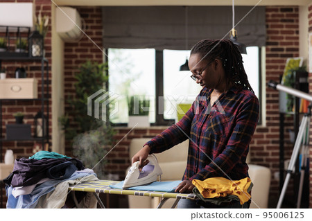 African american woman ironing clothes at home, laundry and ironing day, independent lifestyle in urban apartment, modern housewife doing chores and housework at home. Front view. African american woman ironing clothes at home, laundry and ironing day, independent lifestyle in urban apartment, modern housewife doing chores and housework at home. Front view. 95006125