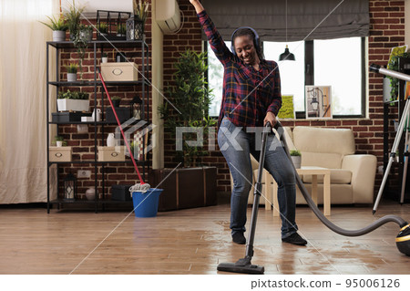 Authentic woman using modern vacuum cleaner listening to favorite music with wireless headphones. Housewife having fun while finishing spring cleaning at home, good vibes and positive energy. 95006126
