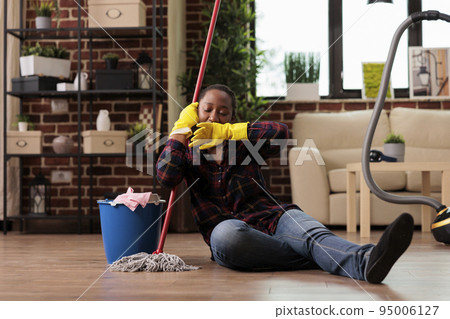 Stressed woman from daily work sitting on the floor with no strength to continue cleaning apartment. Tired housewife holding mop looks away with negative expression of exhaustion. Stressed woman from daily work sitting on the floor with no strength to continue cleaning apartment. Tired housewife holding mop looks away with negative expression of exhaustion. 95006127