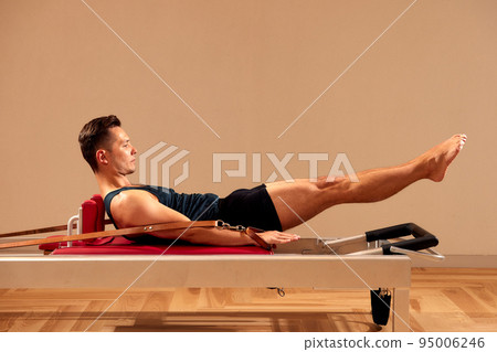 Fit man doing a lunge stretch yoga pilates exercise to strengthen and tone his muscles using a reformer in a gym Fit man doing a lunge stretch yoga pilates exercise to strengthen and tone his muscles using a reformer in a gym 95006246