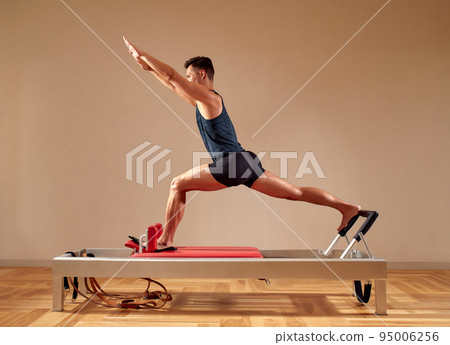 Fit man doing a lunge stretch yoga pilates exercise to strengthen and tone his muscles using a reformer in gym. Health and fitness concept 95006256