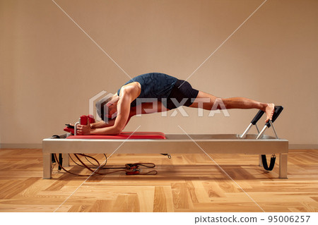 Fit man doing a lunge stretch yoga pilates exercise to strengthen and tone his muscles using a reformer in gym. Health and fitness concept 95006257