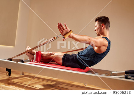 Fit man doing a lunge stretch yoga pilates exercise to strengthen and tone his muscles using a reformer in gym. Health and fitness concept Fit man doing a lunge stretch yoga pilates exercise to strengthen and tone his muscles using a reformer in gym. Health and fitness concept 95006258
