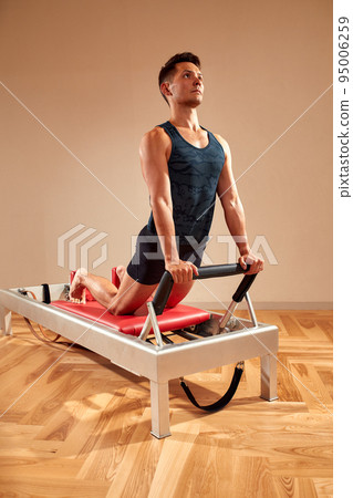 Fit man doing a lunge stretch yoga pilates exercise to strengthen and tone his muscles using a reformer in a gym 95006259