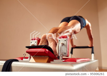 Fit man doing a lunge stretch yoga pilates exercise to strengthen and tone his muscles using a reformer in gym. Health and fitness concept Fit man doing a lunge stretch yoga pilates exercise to strengthen and tone his muscles using a reformer in gym. Health and fitness concept 95006261