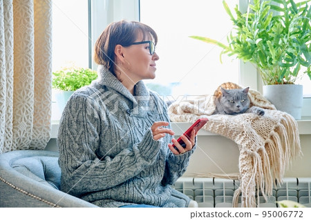 Woman in sweater sitting in armchair near window with heating radiator and sleeping cat Woman in sweater sitting in armchair near window with heating radiator and sleeping cat 95006772