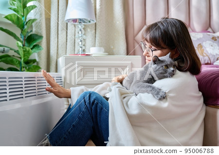 Woman sitting with cat under blanket warming near an electric heater Woman sitting with cat under blanket warming near an electric heater 95006788