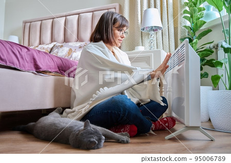 Woman sitting with cat under blanket warming near an electric heater Woman sitting with cat under blanket warming near an electric heater 95006789