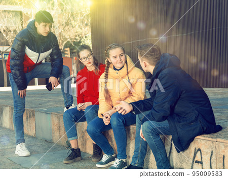 Four teenagers enthusiastically look at the screens of their smartphones on street Four teenagers enthusiastically look at the screens of their smartphones on street 95009583